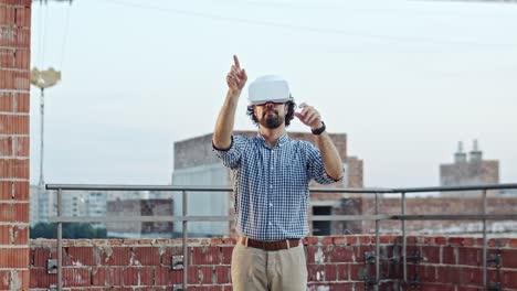 Caucasian-handsome-young-man-architect-or-foreman-in-VR-glasses-standing-on-the-roof-at-the-building-site-and-having-headset-as-watching-virtual-tour-of-future-architecture.-Outdoors.-New-modern-technologies.